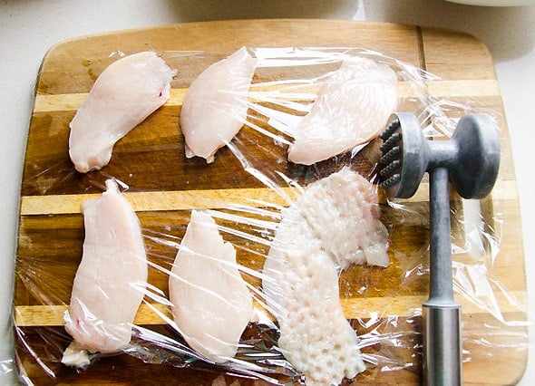 using a meat tenderizer over plastic wrapped chicken on cutting board