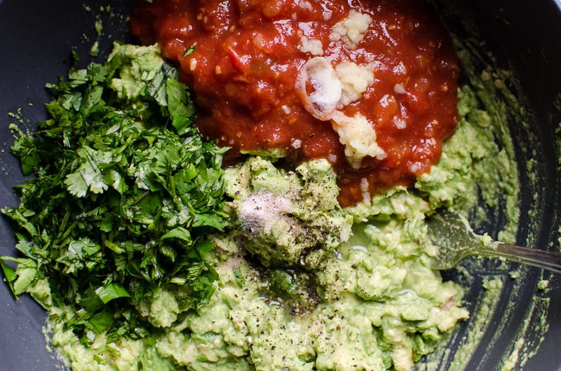 Salsa, mashed avocado, cilantro, garlic, salt and pepper in a bowl with a spoon.