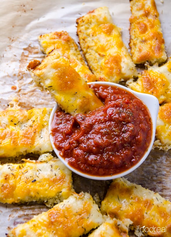 Cauliflower breadsticks on parchment paper with one being dipped into bowl of tomato sauce.