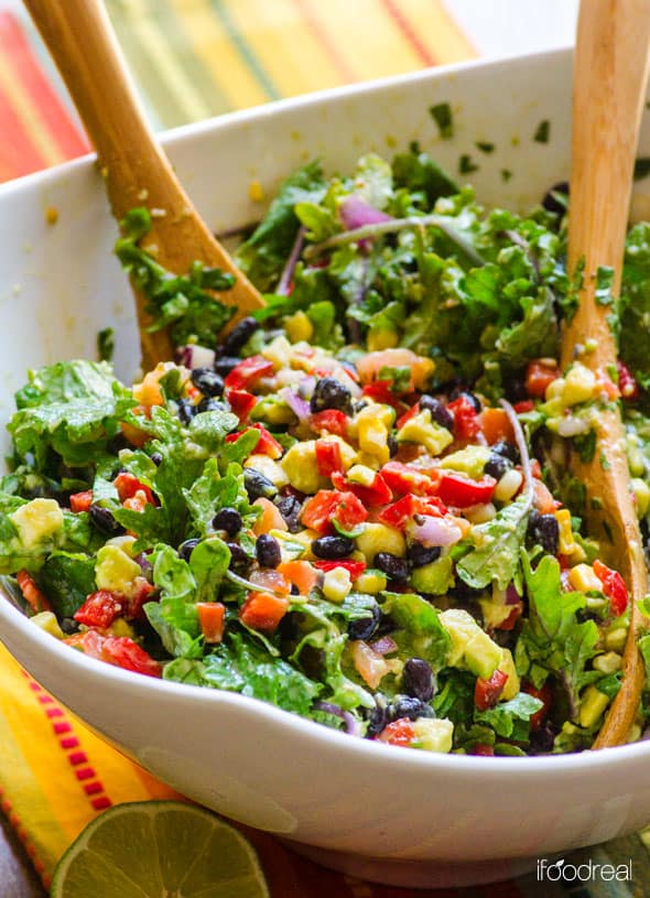 mexican kale salad in a white salad bowl with wooden salad tongs