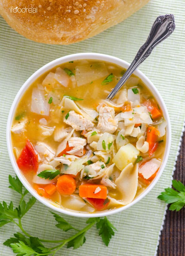 Chicken vegetable noodle soup in a bowl with silver spoon and crusty bread to the back with parsley garnish.