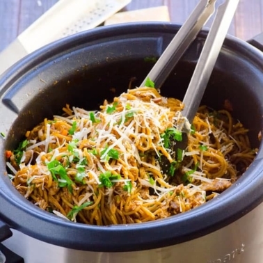 Slow cooker Chicken and Spaghetti with cheese and parsley on top and tongs inside.