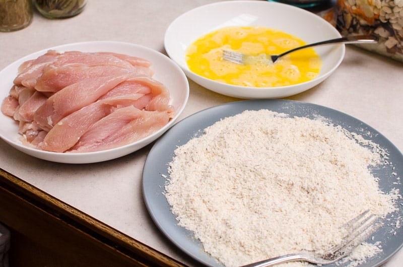 bowl of raw chicken breast, bowl of whisked egg and bowl of coconut flakes, flour and seasoning