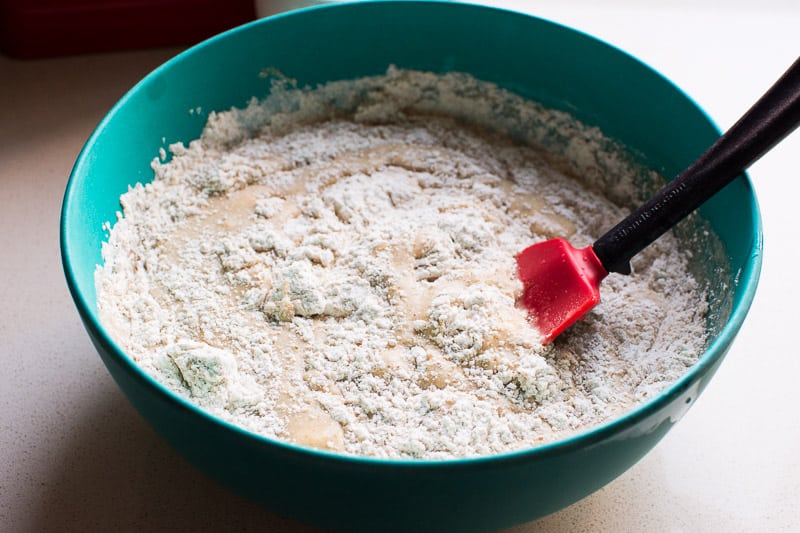 A spatula in a blue bowl stirring flour into liquid ingredients.