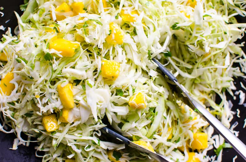 Mango slaw with tongs in black bowl.