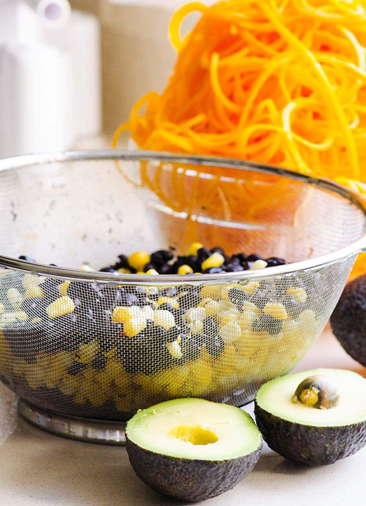Corn and black beans in a colander. SLiced in halves avocado. Butternut squash noodles in a bowl.