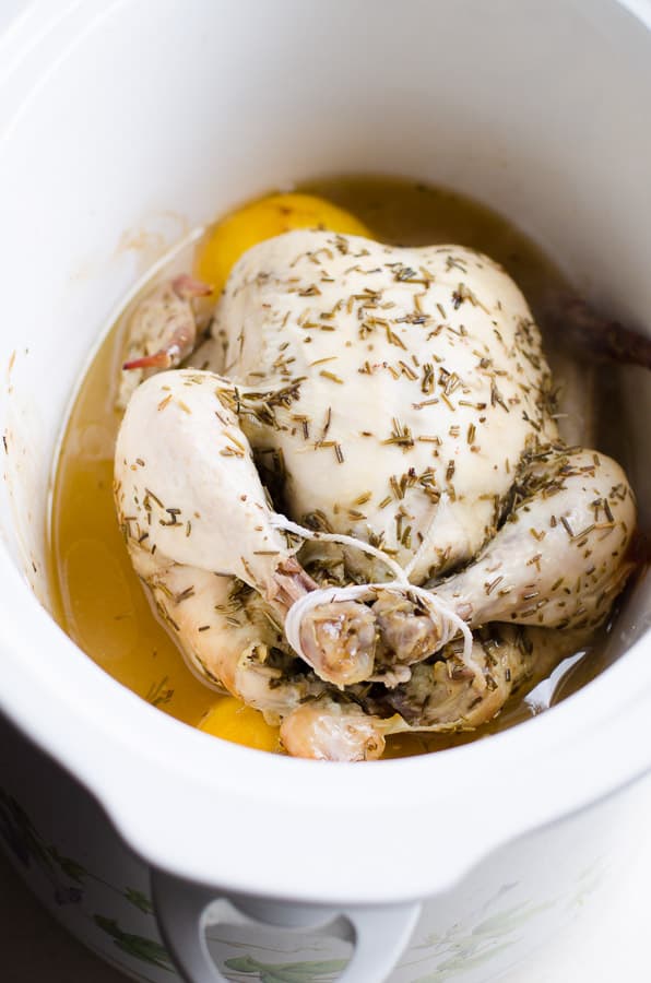 Cooked whole chicken seasoned with spices in slow cooker with broth and lemons.