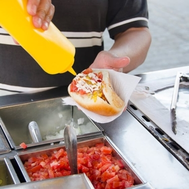 A person holding a hot dog and adding mustard on it.
