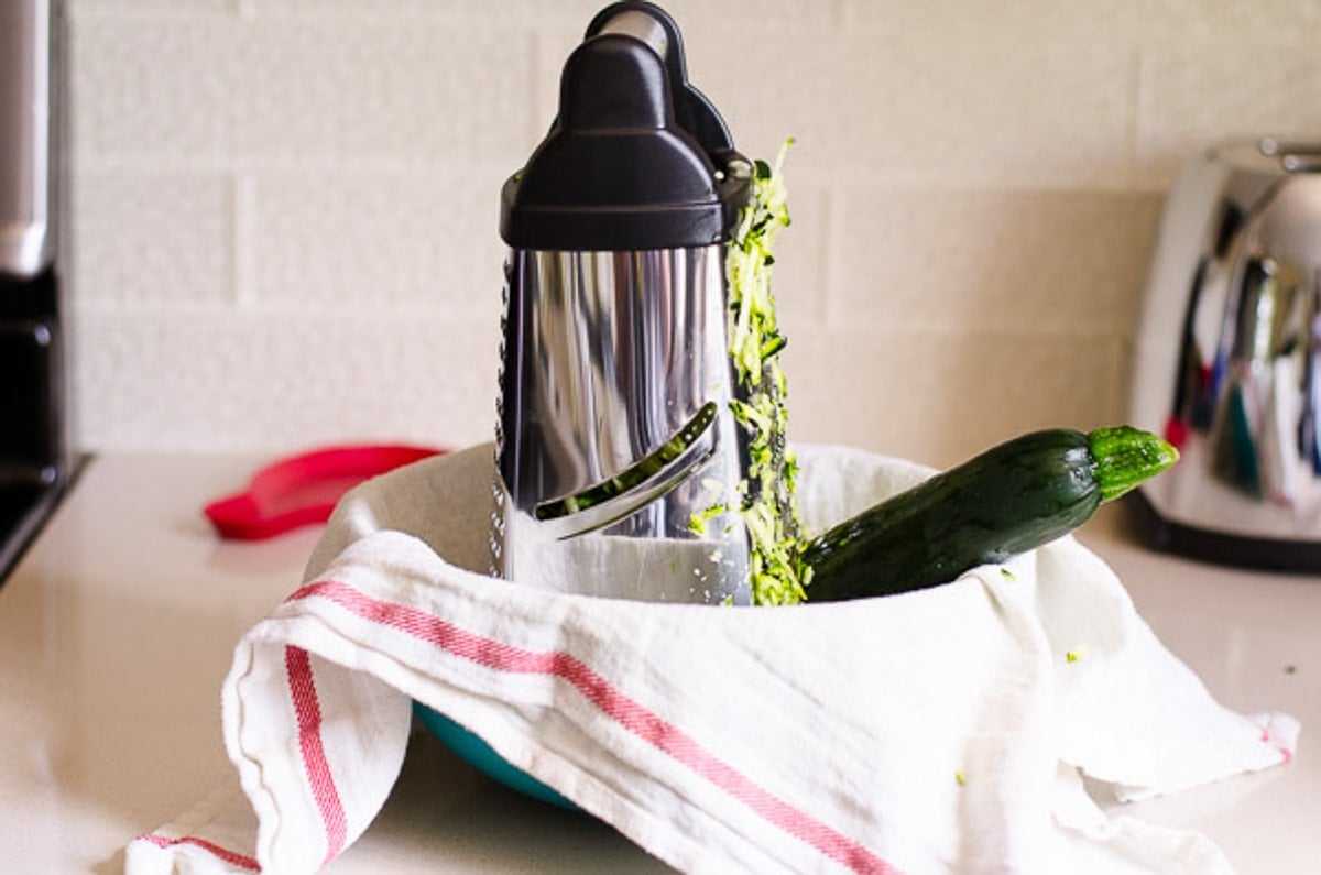 Box grater with a zucchini in a bowl lined with linen towel. 