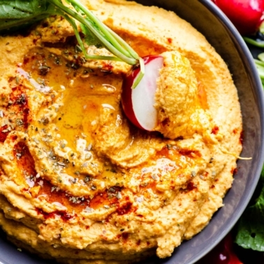 Sweet potato hummus drizzled with oil and served in a bowl with vegetables around it.