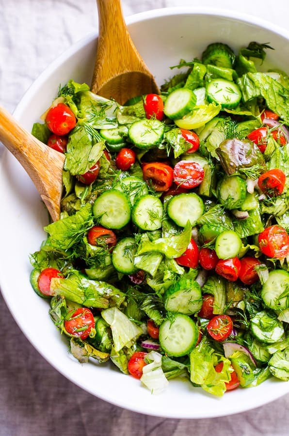 Lettuce salad with tomato and cucumber and fresh dill in white serving bowl with wooden spoons.