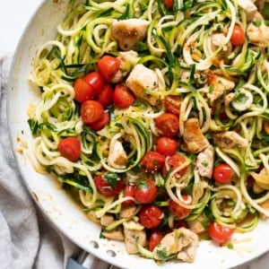 Zucchini noodles with chicken and grape tomatoes in white skillet and linen napkin nearby.
