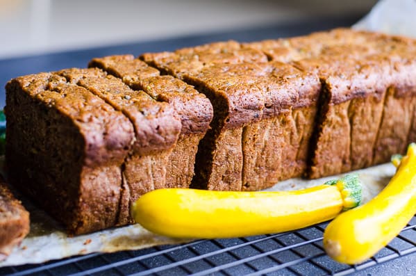 Sliced zucchini bread on cooling rack and two yellow squash nearby.