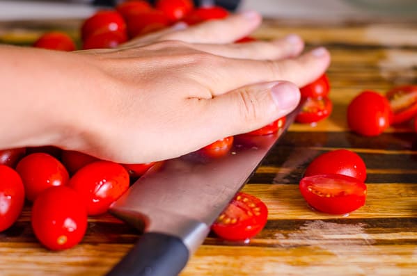 Person pressing on grape tomatoes with one hand and slicing through them with a knife on a cutting board.
