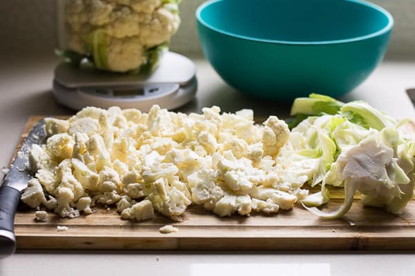 Chopped cauliflower with leaves separated on a cutting board with a knife; bowl and scales behind.