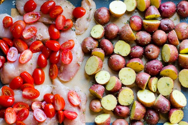 Chicken topped with cherry tomatoes and potatoes on baking sheet.