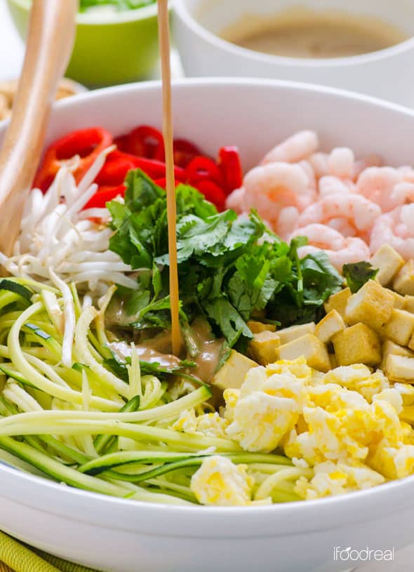 Pouring peanut sauce over zucchini noodles, shrimp, eggs, tofu, bean sprouts and red pepper in a bowl.