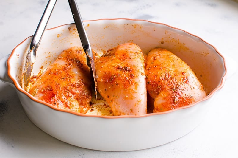 Seasoned and tossed with tongs three raw chicken breasts in a baking dish.