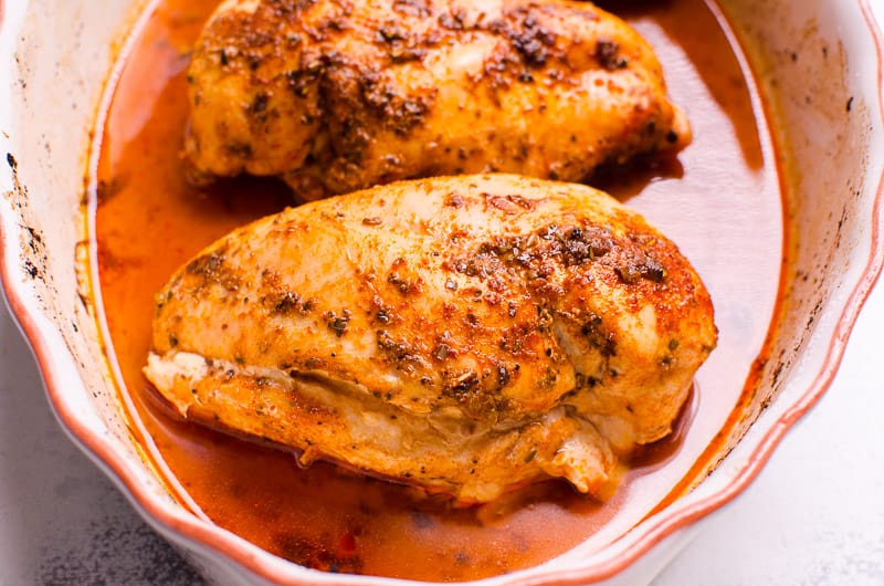 two baked chicken breasts with juices in white baking dish