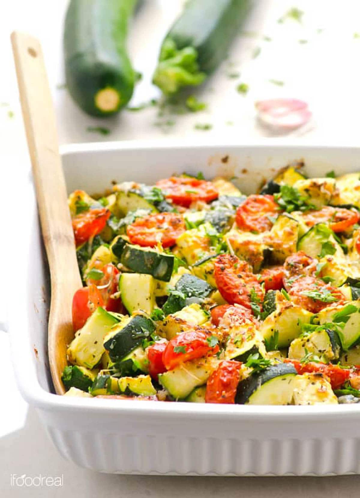 Zucchini tomato bake in white baking dish with wooden spoon.