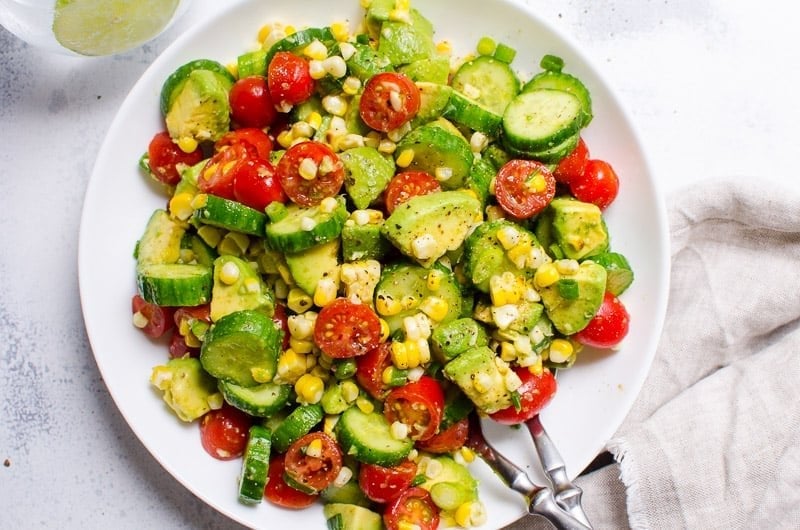 Avocado corn salad cucumbers and tomatoes in a white bowl with serving utensils.