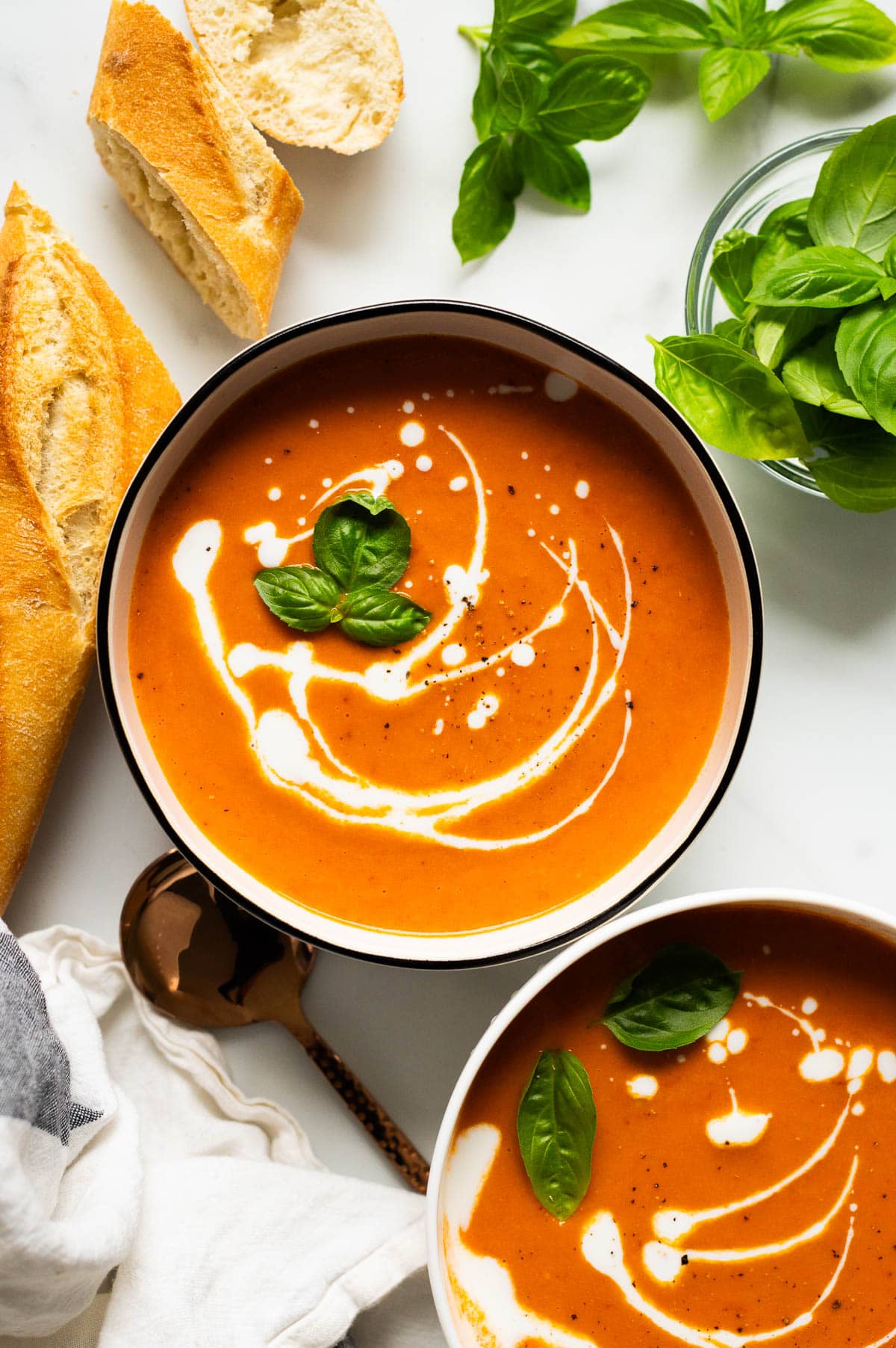 Creamy tomato soup served in bowls and garnished with cream and basil. Bread and basil on a counter.