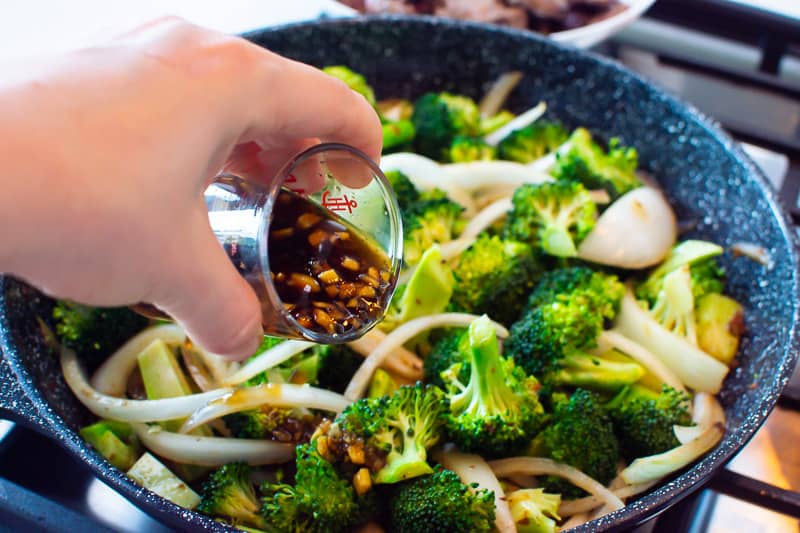 Sauce in small measuring jar being added to broccoli in skillet.
