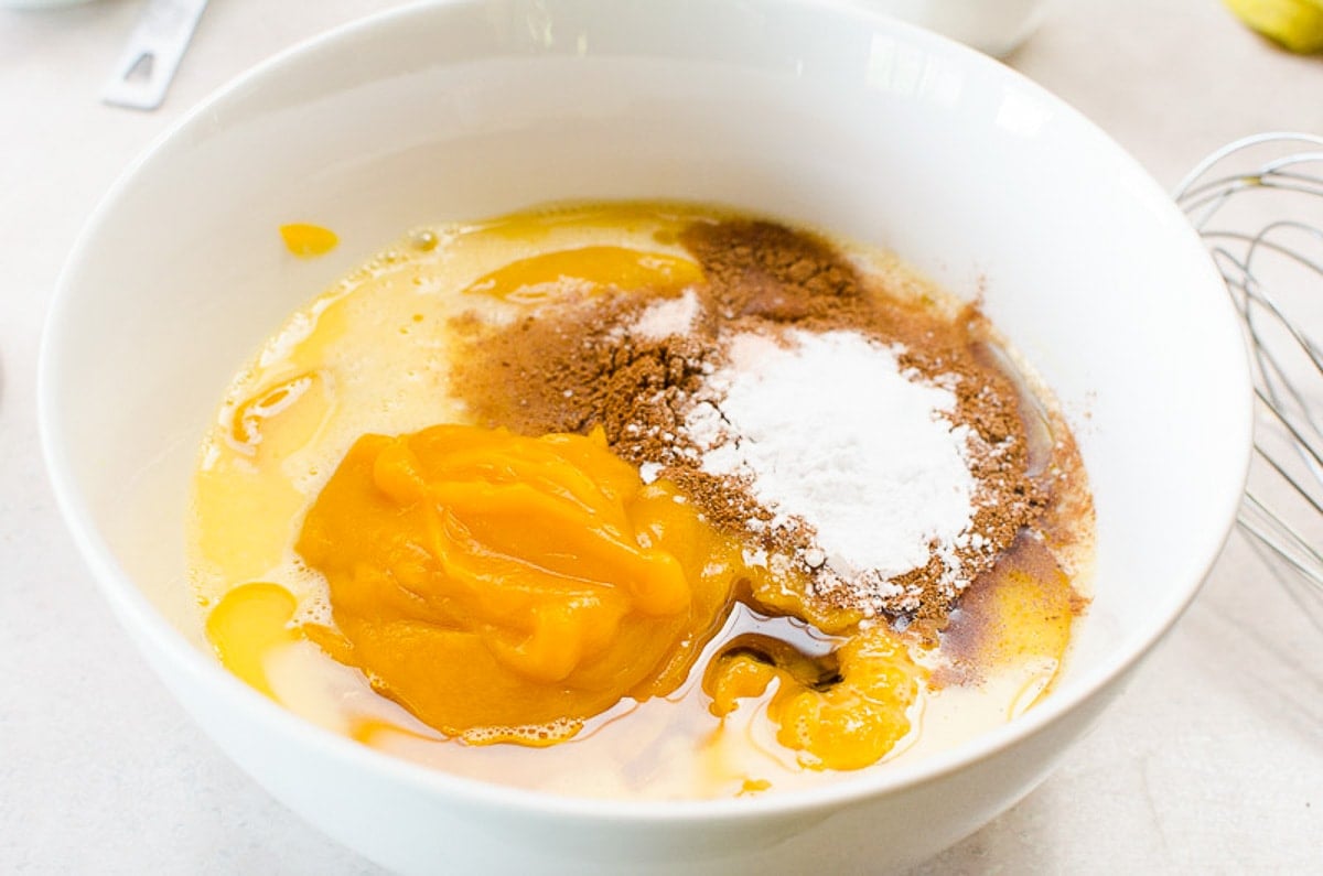 Pumpkin, egg, maple syrup, cinnamon and baking staples in white bowl.