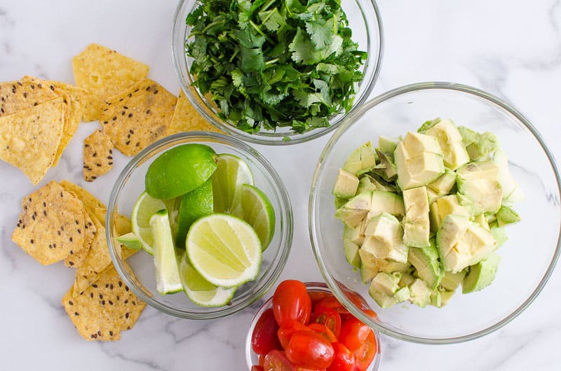 Chopped avocado, cilantro, tomatoes, limes in separate bowls and tortilla chips.