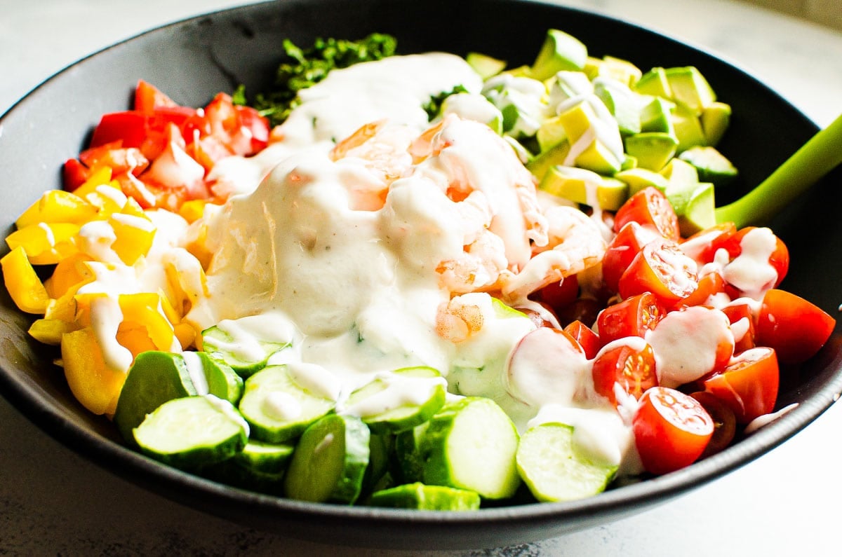 Shrimp, tomatoes, cucumber, peppers, avocado and cilantro with yogurt dressing in a bowl.