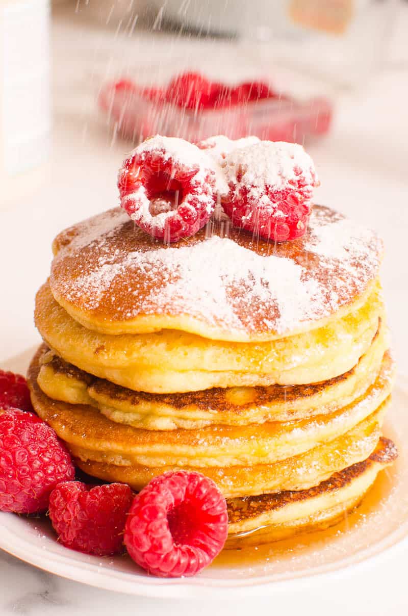 White plate of almond flour pancakes topped with raspberries and icing sugar.