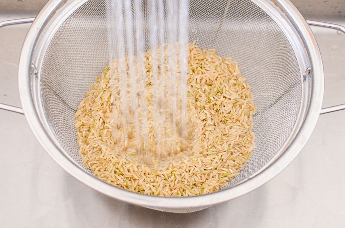 Brown rice in colander being rinsed with water.