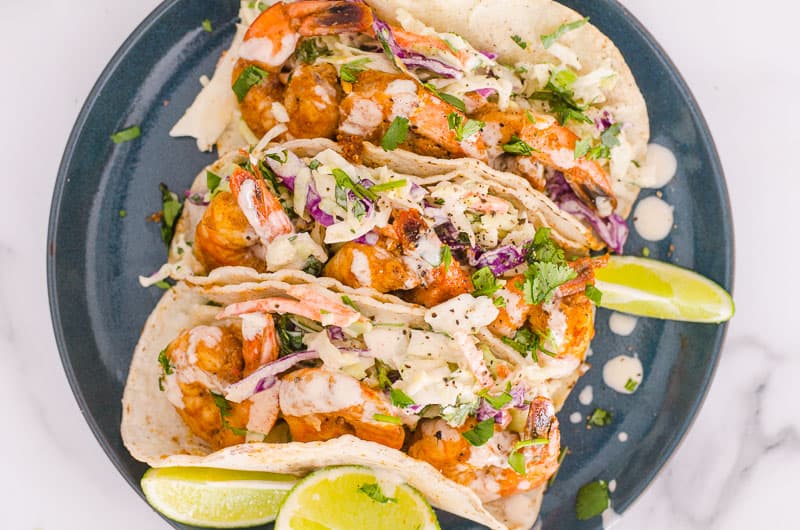 Three shrimp tacos on plate for serving with lime wedges.