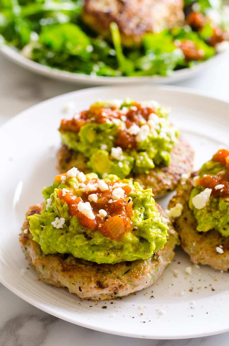 Three turkey burgers on a plate topped with guacamole, salsa and cheese.
