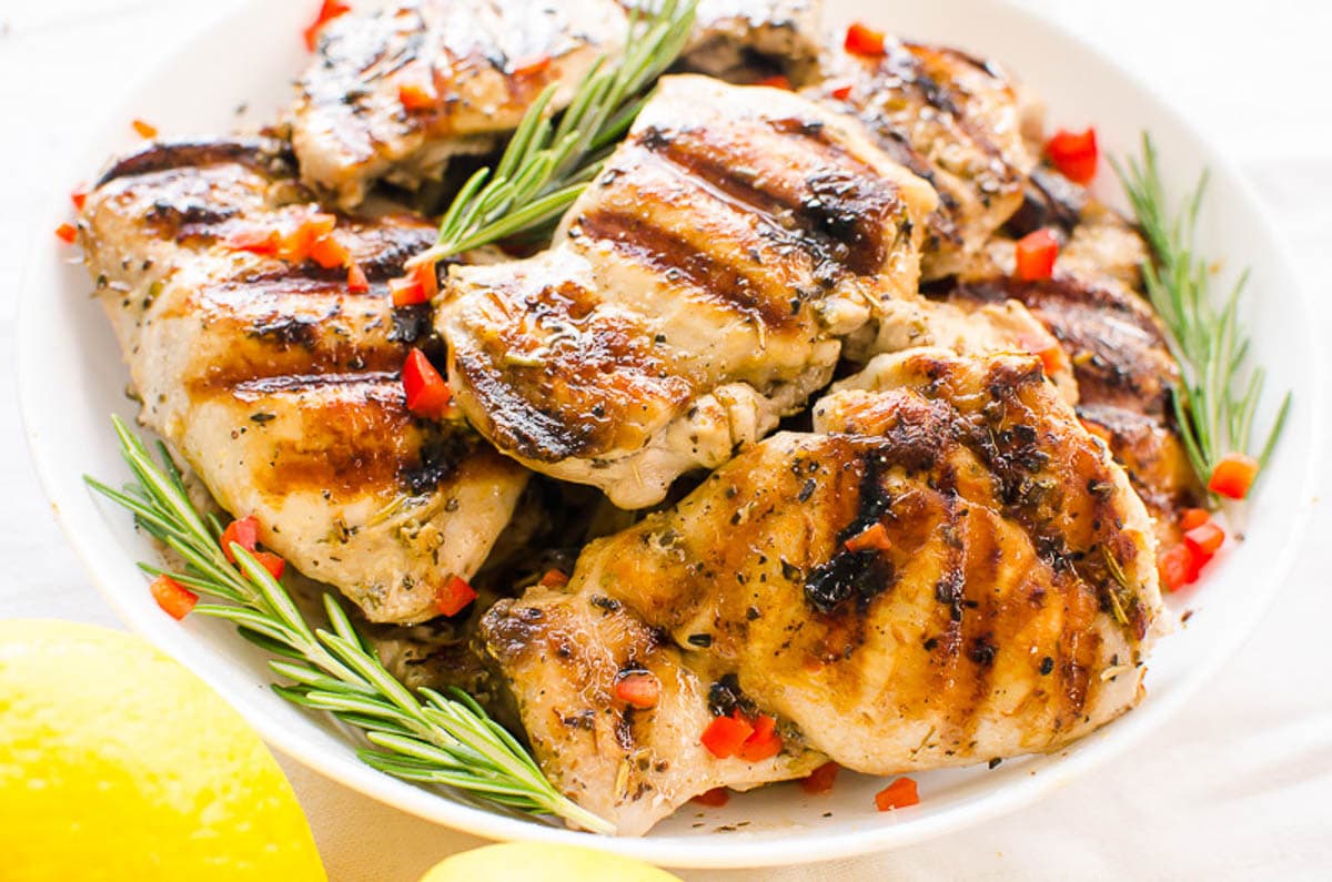Grilled Greek chicken thighs garnished with pepper and rosemary on white plate.