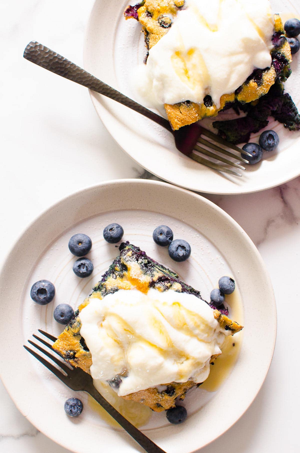 Two slices of blueberry breakfast cake on plates with yogurt and fresh blueberries.