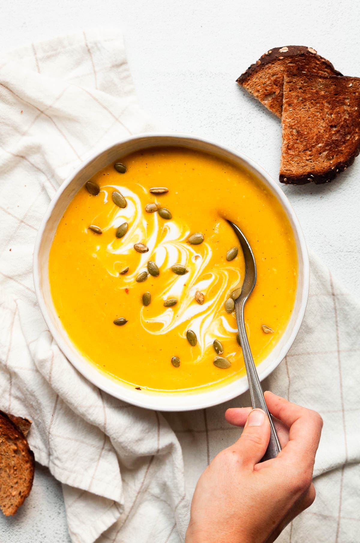 Person dipping a spoon into a bowl with butternut squash soup garnished with cream and pumpkin seeds.