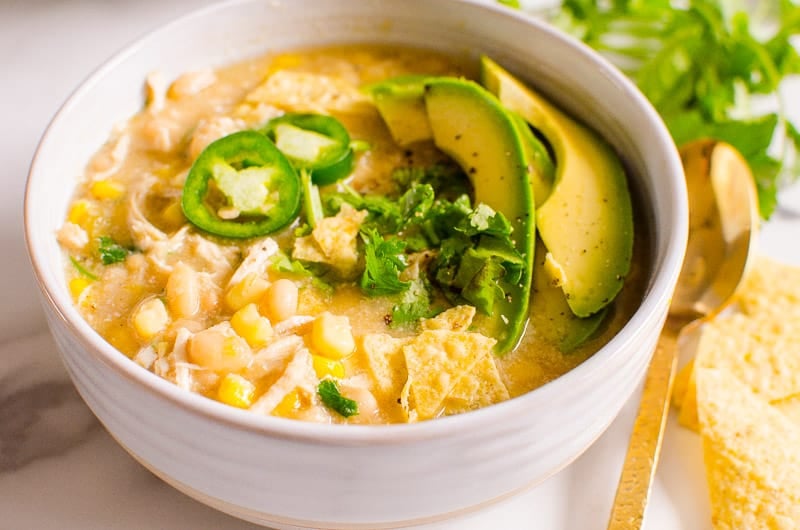 White bean chicken chili garnished with fresh cilantro, avocado and jalapenos in a bowl. Spoon, tortilla chips and cilantro on a counter.