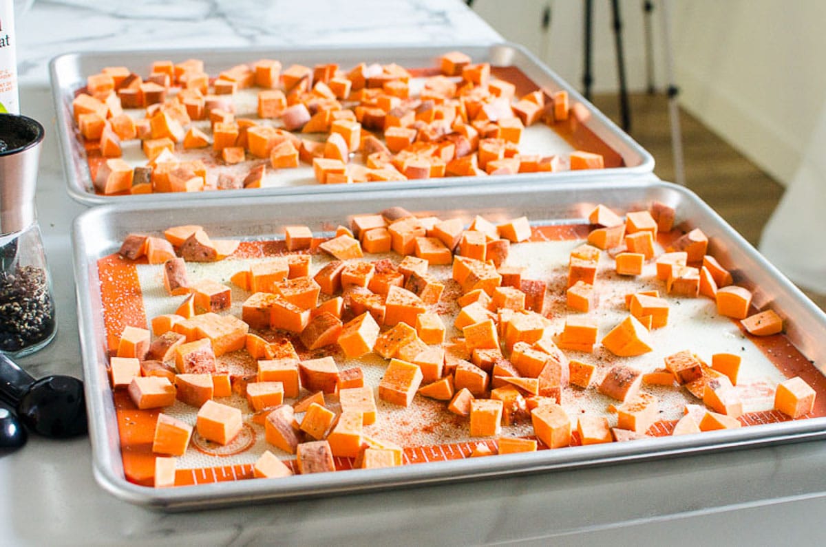 Two baking sheets with uncooked seasoned diced sweet potatoes.