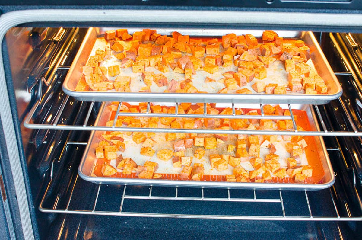 Two sheet pans of cubed sweet potatoes in the oven with lights on.