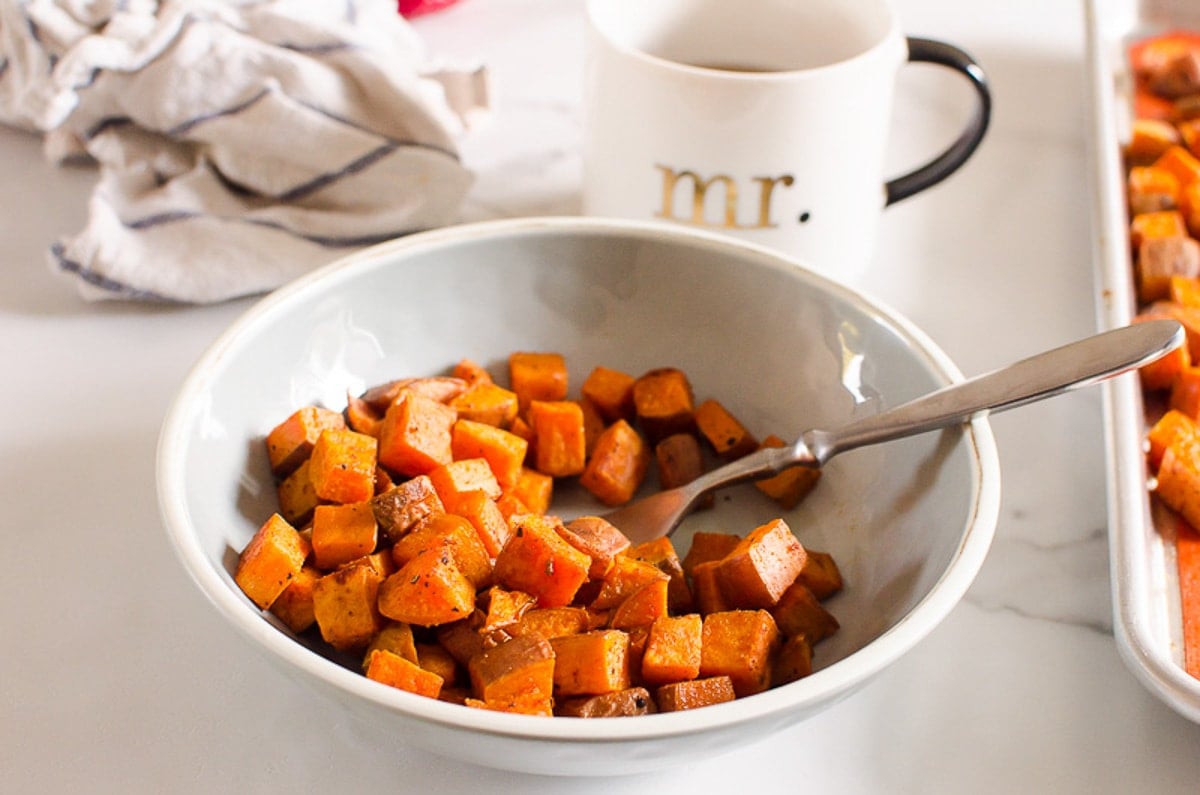 A bowl with roasted sweet potatoes and fork. A mug with tea, towel and baking sheet with more food.