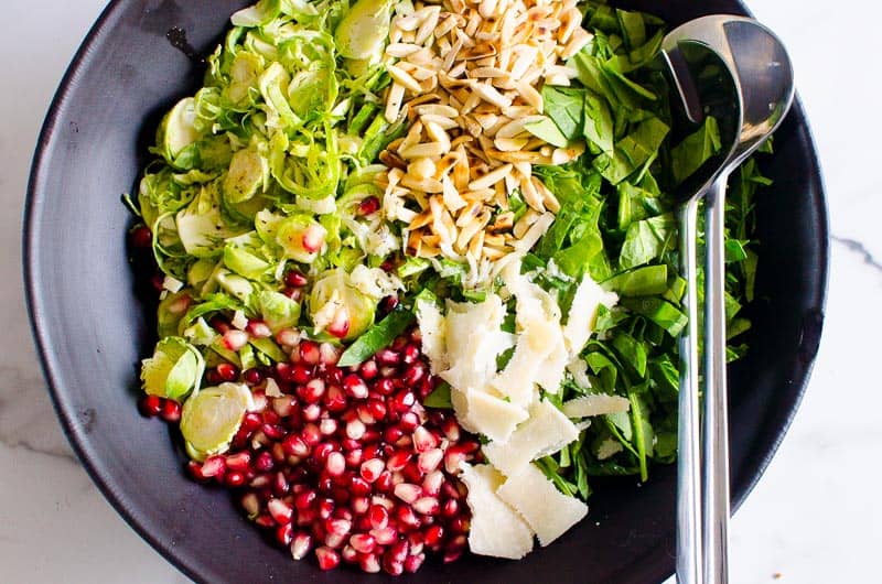 Shaved Brussels sprouts, pomegranate arils, slivered almonds and shaved Parmesan cheese in black bowl with metal spoons. 