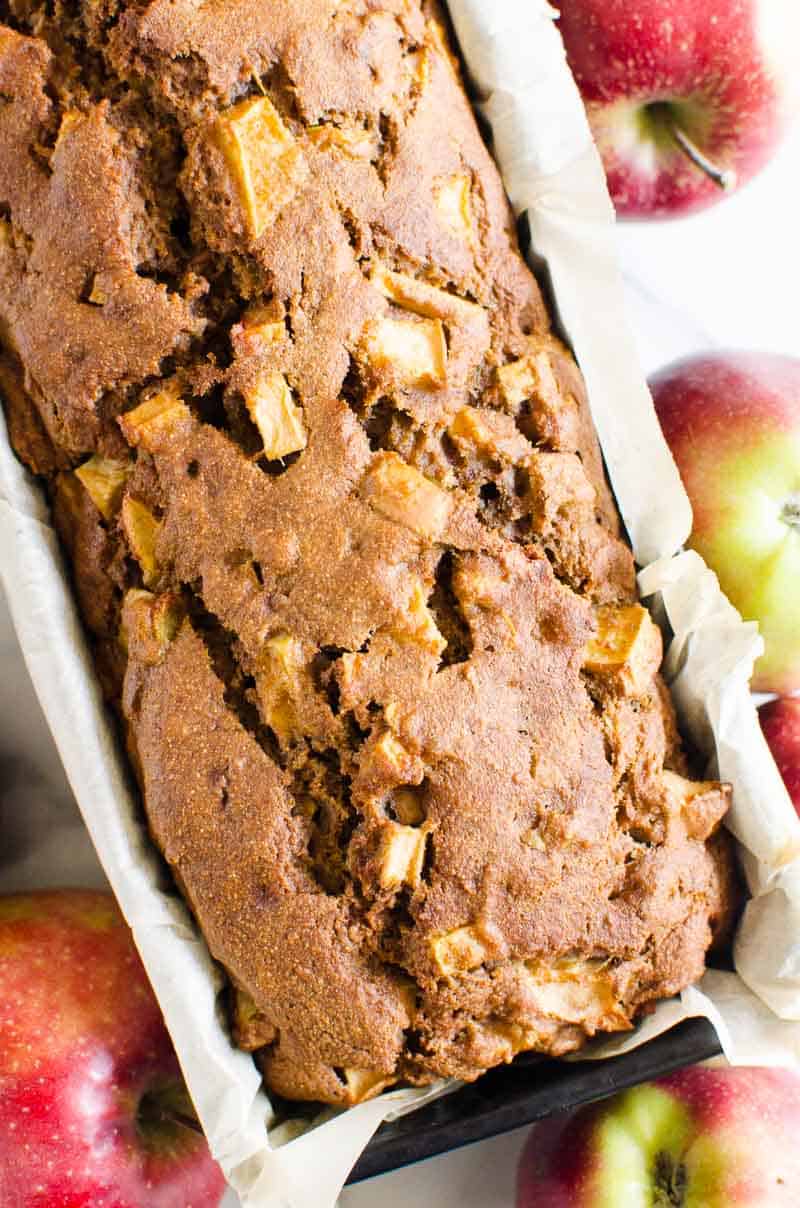 Healthy apple bread in a parchment lined loaf pan with fresh apples beside the pan. 