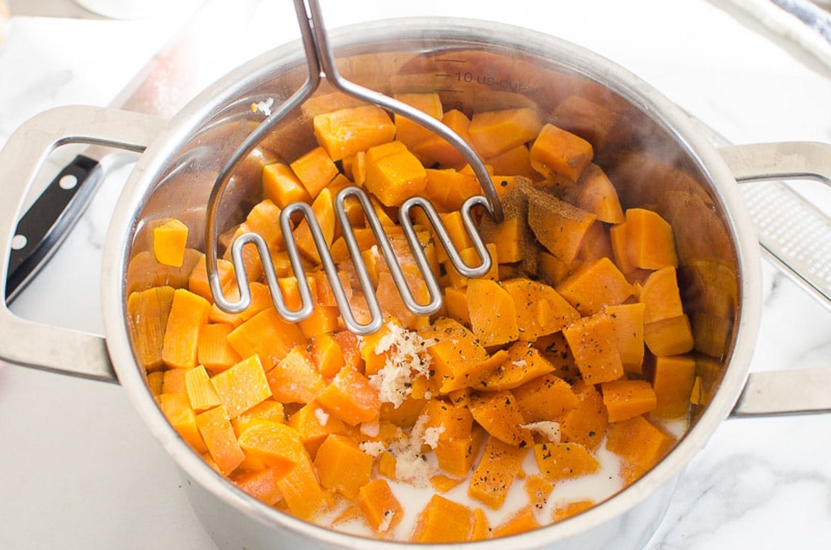Mashing cooked sweet potatoes with milk and spices in a stainless steel pot.
