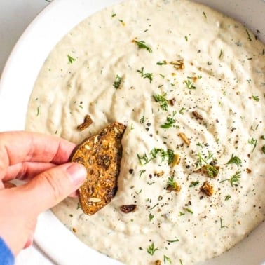 Healthy french onion dip in a bowl with a cracker being dipped into it.
