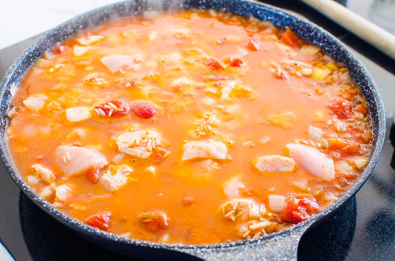 Chicken and rice ingredients simmering in a broth in a skillet. 