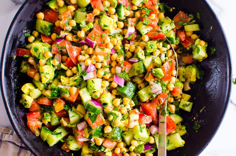 Chickpea salad with avocado, parsley, tomato, cucumber and red onion in black bowl.