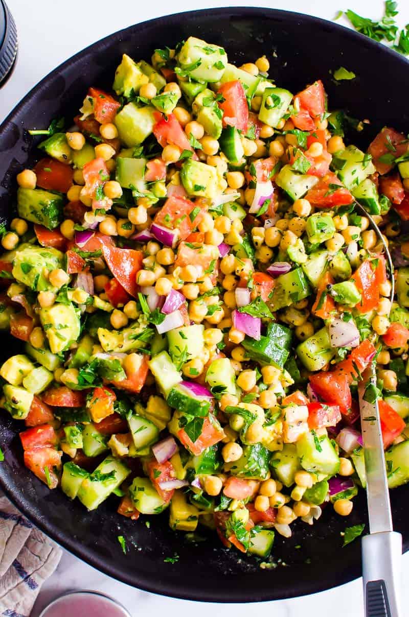 Chickpea salad with avocado in black bowl with metal spoon.