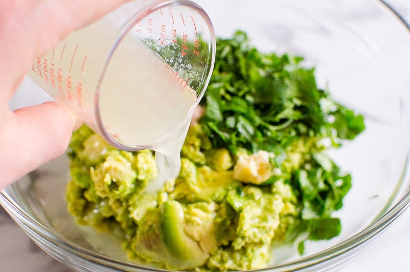 Pouring lime juice over mashed avocado and cilantro in a bowl.