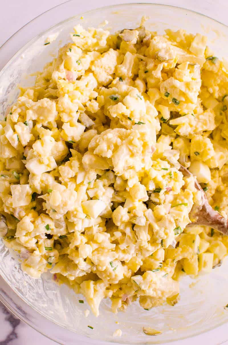 Cauliflower potato salad in glass bowl with wooden spoon.